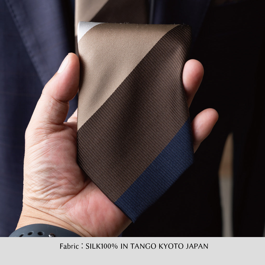 手に持ったワイドストライプのシルクネクタイ、ブラウン×ネイビー系